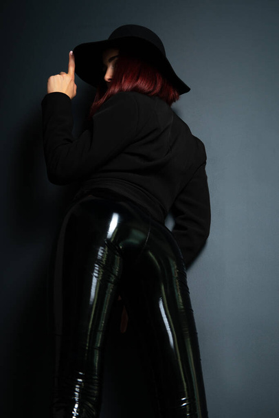 Beautiful fashion woman with perfect body, back view. Portrait of beautiful girl wearing black blazer, latex tight pants and hat posing in studio over deep  gray background - Fotoğraf, Görsel
