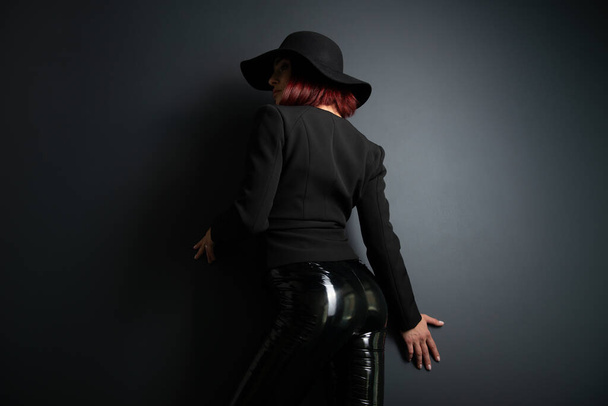 Beautiful fashion woman with perfect body, back view. Portrait of beautiful girl wearing black blazer, latex tight pants and hat posing in studio over deep  gray background - Foto, immagini