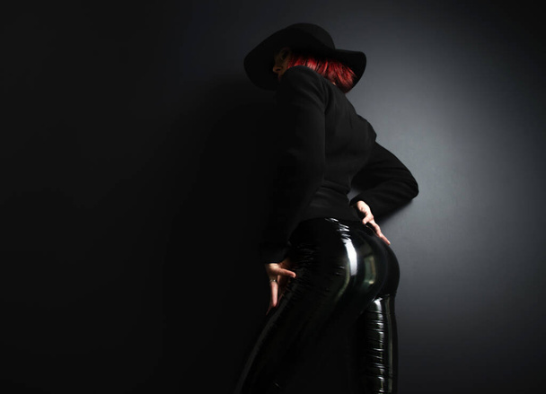 Beautiful fashion woman with perfect body, back view. Portrait of beautiful girl wearing black blazer, latex tight pants and hat posing in studio over deep  gray background - Foto, Bild
