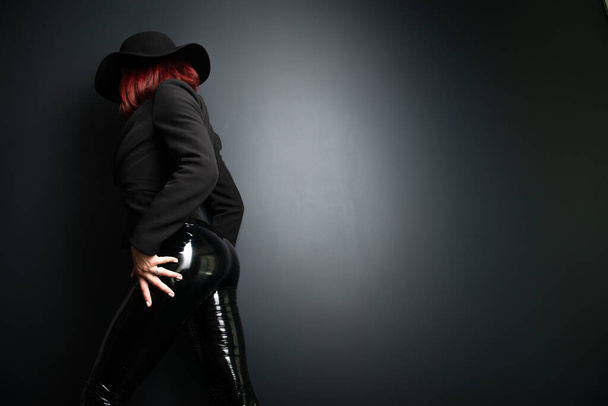 Beautiful fashion woman with perfect body, back view. Portrait of beautiful girl wearing black blazer, latex tight pants and hat posing in studio over deep  gray background - Valokuva, kuva
