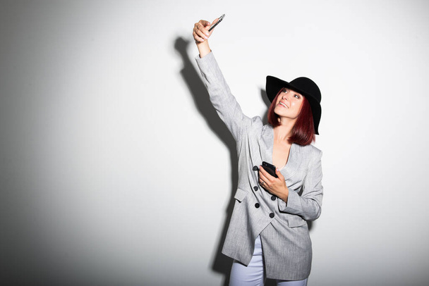 Young stylish woman using smartphone, indoor portrait over white. Fashion modern girl wearing grey jaket and black hat taking selfie photo with mobile phone - Foto, imagen