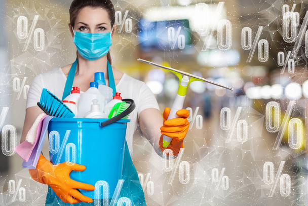 Una donna delle pulizie in maschera sta con gli strumenti di pulizia sullo sfondo di sconti percentuali di volo. - Foto, immagini