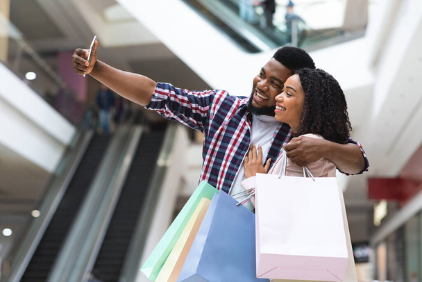 Cónyuges negros positivos comprando en grandes almacenes y tomando selfie en el teléfono inteligente - Foto, Imagen