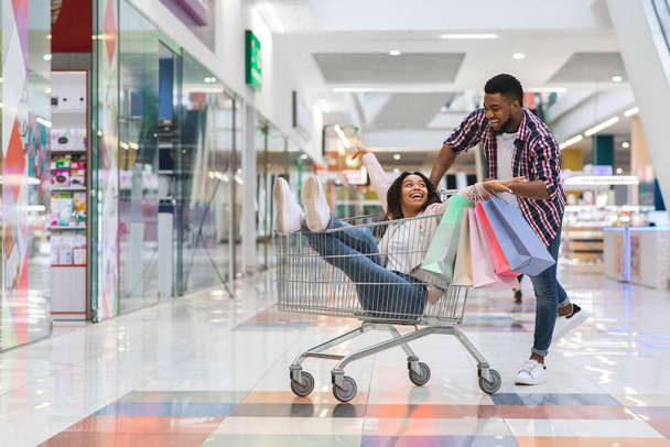 alegre negro hombre tirando de carro de compras con su novia dentro, divertirse - Foto, imagen