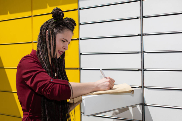 A young girl is addressing the letter while standing next to the parcel locker. Use a ballpoint pen to write on the gray letter envelope. Attractive teenage girl. - Foto, imagen