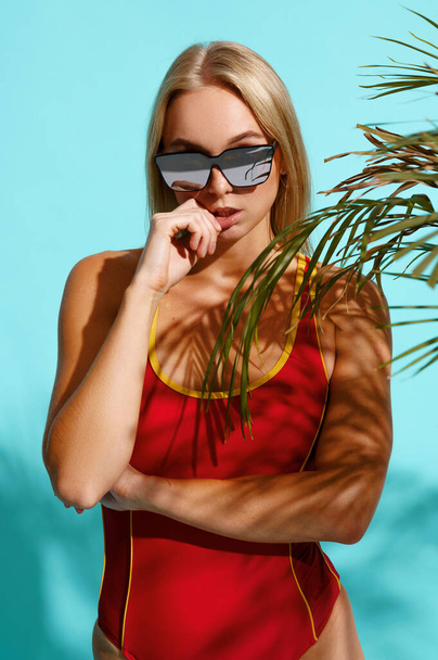 Kırmızı mayo giymiş sportif bir kadın stüdyoda Cyan arka planda poz veriyor. Mayolu kız bronzlaşmaya hazır. Yüzme iç çamaşırlı, ince vücutlu, baştan çıkarıcı kadın yüzücü. - Fotoğraf, Görsel