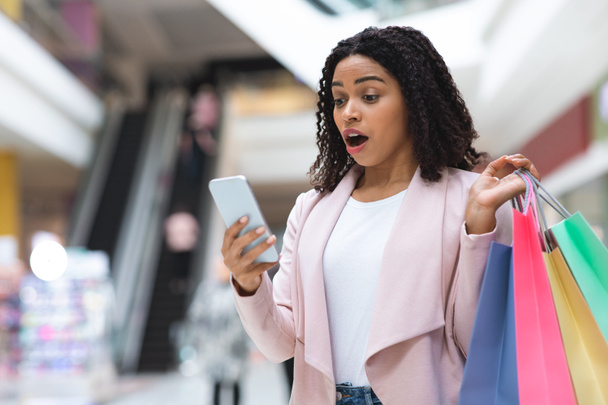 Ritratto di donna nera scioccata con borse della spesa e smartphone nel centro commerciale - Foto, immagini