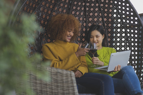 Két nő pihen a hintában, a legjobb barátok vörösbort isznak. Multiracionális kapcsolat vagy homoszexuális kapcsolat - Fotó, kép