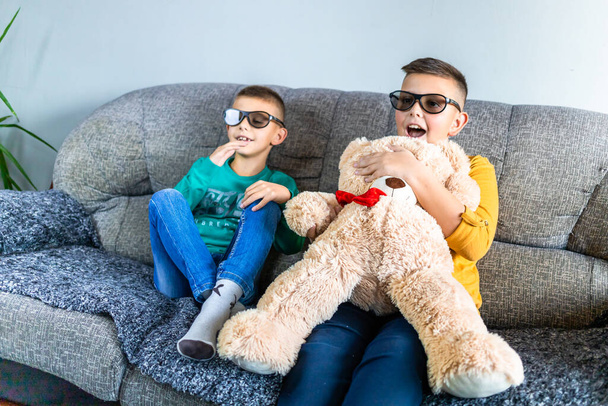 Cute boys, bothers watching some scary movie with 3d glasses, sitting on the sofa in living room, together with teddy bear toy - Photo, image
