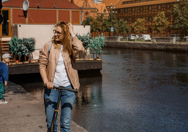 Beautiful girl with glasses. Young woman with a scooter in the city. Pretty girl by the river in the city. Journey to Bydgoszcz. - Фото, зображення