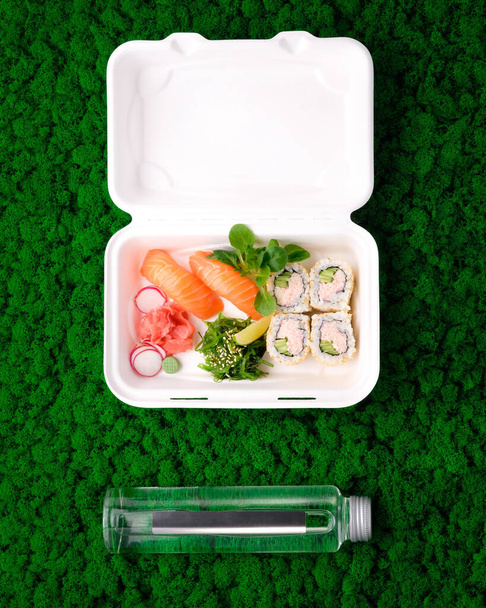 Sushi set in takeout box with water in glass bottle on green moss background. Salmon sashimi, California maki roll. Takeaway japanese food reastaurant concept. Eco-friendly, sustainable packaging. - Photo, Image