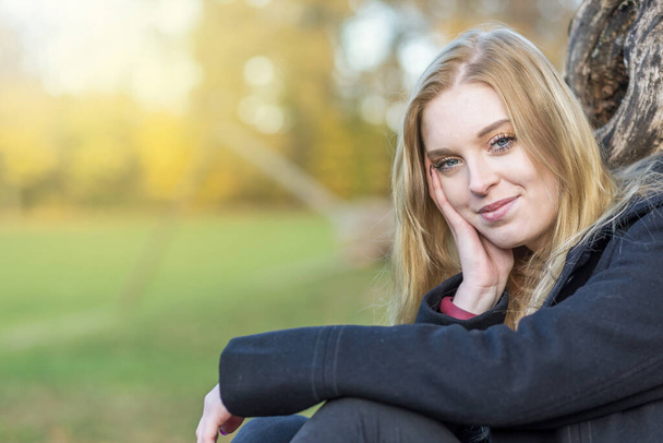 Smiling young woman is posing in the autumn park supporting her head with  hand looking at the camera. Horizontally.  - Foto, imagen