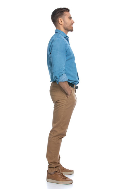 πλαϊνή άποψη ενός casual νεαρός άνδρας στέκεται με τα χέρια του στις τσέπες του και κοιτάζοντας μπροστά με ένα χαμόγελο στο πρόσωπό του σε λευκό φόντο - Φωτογραφία, εικόνα