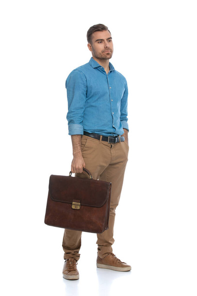 young casual man looking away, holding his briefcase and sticking one hand in his pocket against white background - Photo, Image