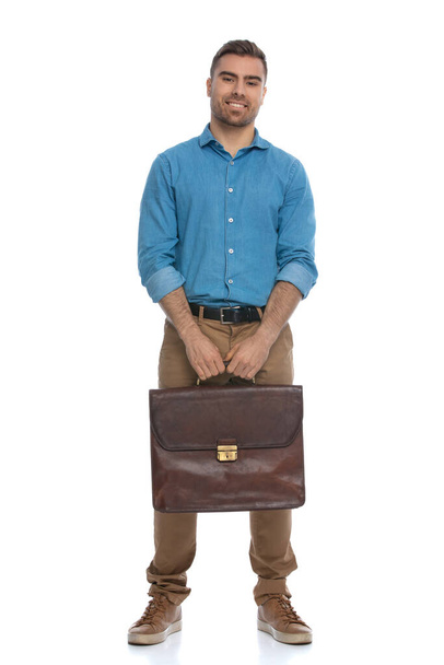 sexy hombre casual sosteniendo un maletín en sus manos y sonriendo a la cámara sobre fondo blanco - Foto, imagen