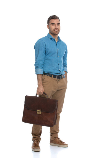 guapo hombre casual sosteniendo su maletín con una mano y metiendo la otra en su bolsillo contra fondo blanco - Foto, Imagen