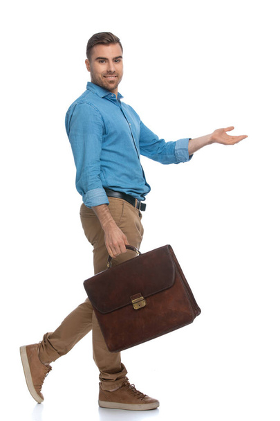 joven hombre casual caminando y señalando a un lado, mientras mira a la cámara y sostiene un maletín sobre fondo blanco - Foto, Imagen