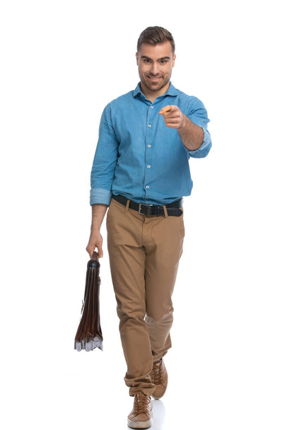 hombre casual guapo apuntando a la cámara, caminando y sosteniendo un maletín sobre fondo blanco - Foto, Imagen