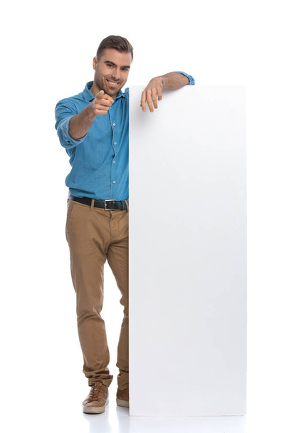 casual young man pointing at the camera while leaning on a long billboard against white background - Photo, Image