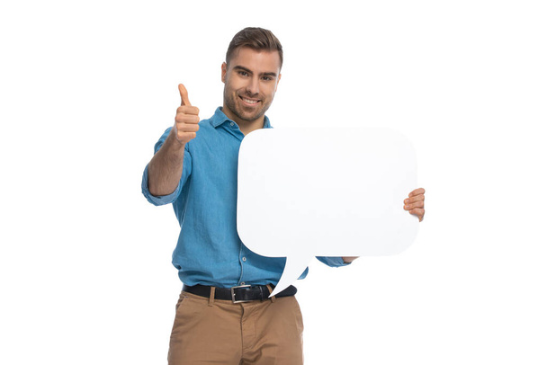 attractive casual man holding a speech bubble and giving a thumbs up against white background - Foto, afbeelding
