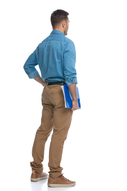 vista trasera de un joven hombre casual sosteniendo un portapapeles azul y sopesando algo sobre fondo blanco - Foto, imagen