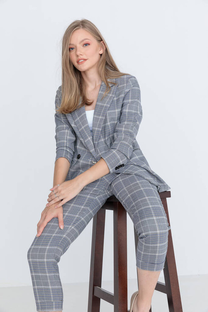 heureux modèle en costume à carreaux gris tenant la main sur les cuisses dans une pose de mode, souriant et assis sur fond gris clair en studio - Photo, image