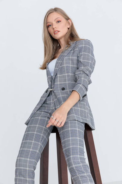 cool blonde model in gray checkered suit looking down and confidently posing, sitting on light gray background in studio - Zdjęcie, obraz