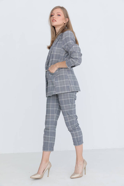 cool fashion model wearing gray checkered suit holding hand in pocket, walking and posing in a side view position on light gray background in studio - Photo, image