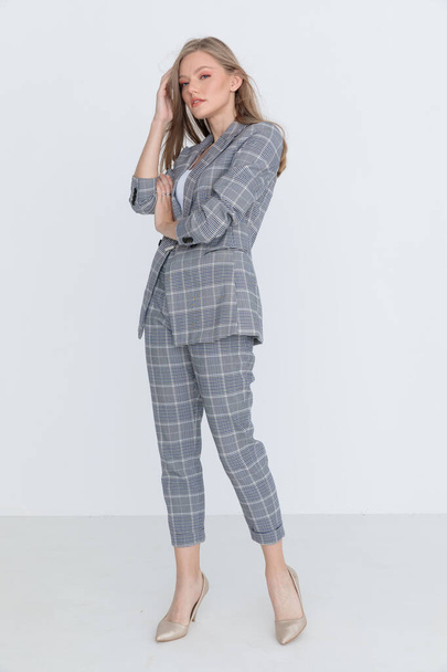 confident businesswoman in gray checkered suit holding hand to hair and arranging, holding arm in a fashion pose and standing on light gray background in studio - 写真・画像