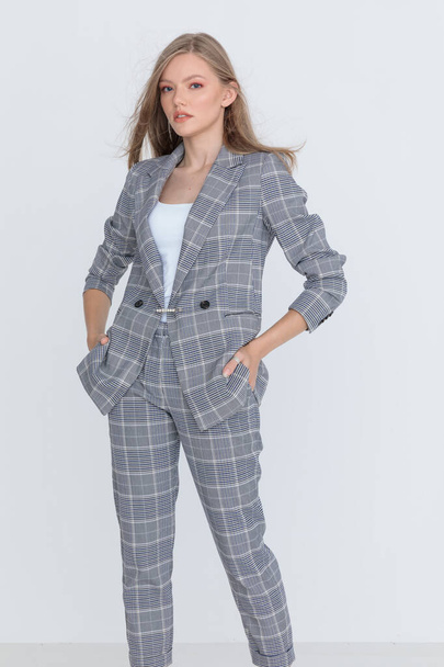 sexy young woman in checkered suit holding hands in pockets, confidently posing on light gray background in studio - Foto, Imagem