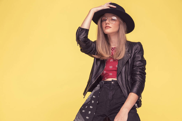 beautiful fashion model adjusting her hat, wearing a cool leather jacket and glasses and sitting against yellow background - Photo, Image