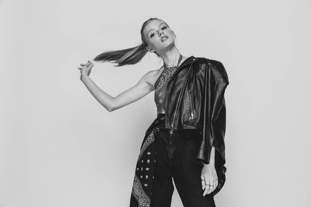 casual hippie girl wearing leather jacket on shoulder, flipping ponytail and posing on black and white background in studio - Photo, Image