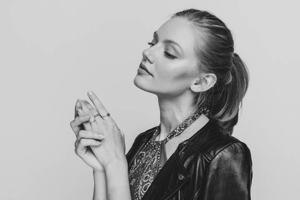 πλαϊνή άποψη της νεαρής γυναίκας με δερμάτινο μπουφάν κοιτάζοντας προς τα πλάγια, αγγίζοντας τα χέρια και θέτοντας σε μαύρο και άσπρο φόντο στο στούντιο - Φωτογραφία, εικόνα