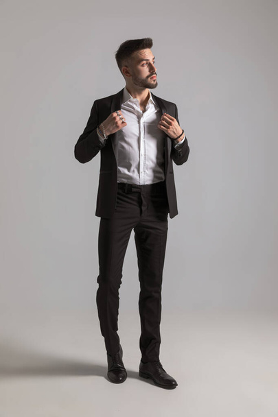 attraktives Geschäftsmann-Modell, das zur Seite schaut und Smoking anpasst, geht und posiert auf hellgrauem Hintergrund im Studio - Foto, Bild