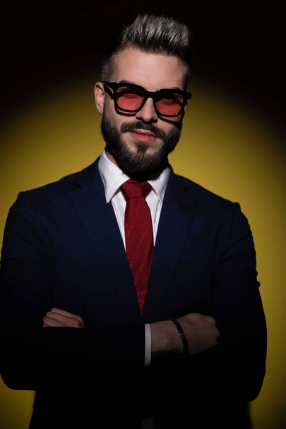 happy young businessman in navy blue suit crossing arms, posing and smiling, standing in a fashion light on yellow background in studio - Zdjęcie, obraz