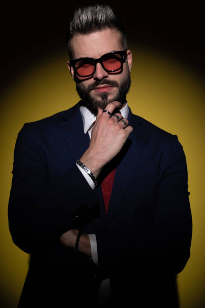 thoughtful young businessman in navy blue suit holding hand to chin and wondering while posing in a fashion light on yellow background in studio - Photo, Image
