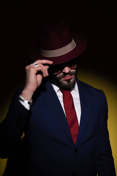 dramatic elegant man in navy blue suit pulling hat over face and hiding, looking down and posing in a fashion light on yellow background - Photo, Image