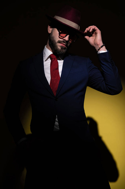 unrasierter junger Geschäftsmann im marineblauen Anzug, die Hände in den Taschen und den Hut arrangierend, posierend im Modelicht auf gelbem Hintergrund - Foto, Bild