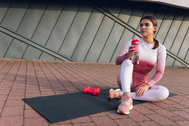Fitness tyttö istuu pullon vettä matolla. Naisurheilija lepää harjoittelun jälkeen - Valokuva, kuva