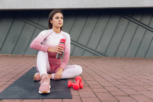 Fitness tyttö istuu pullon vettä matolla. Naisurheilija lepää harjoittelun jälkeen - Valokuva, kuva