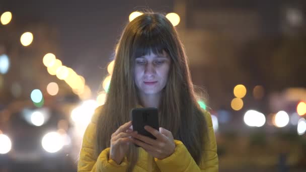 Jovem mulher bonita usando seu telefone celular em pé na rua da cidade à noite ao ar livre. - Filmagem, Vídeo