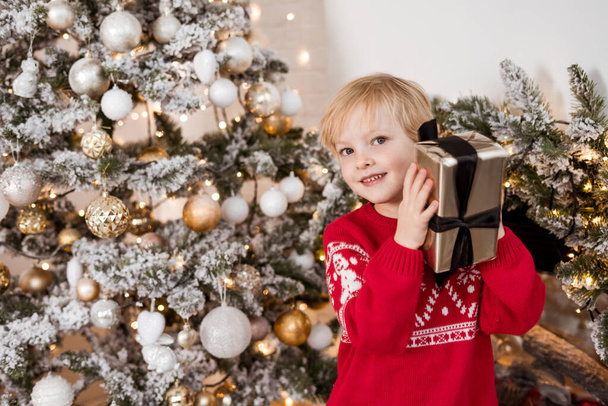 Schauen Sie, welches Geschenk ich zu Weihnachten bekommen habe. glücklicher Junge, roter Pullover, Geschenkbox hochhaltend, in die Kamera blickend und lächelnd, im gemütlichen Wohnzimmer in der Nähe des Weihnachtsbaums stehend - Foto, Bild