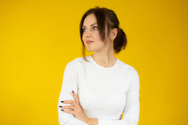 Smiling young brunette woman in white sweater posing isolated on yellow background studio portrait. People sincere emotions lifestyle concept. Mock up copy space. Holding hands crossed - Fotó, kép