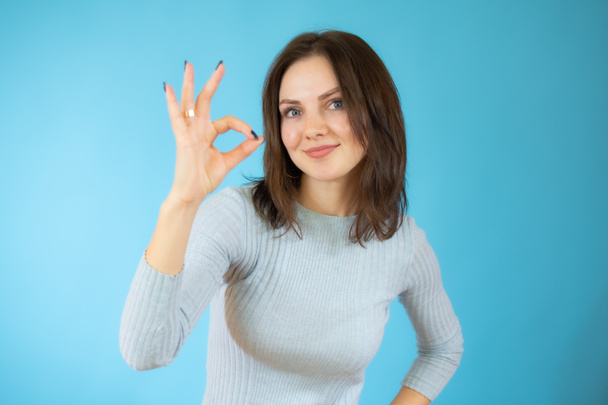 Young beautiful asian woman wearing casual sweater standing over blue isolated background smiling positive doing okay sign with hand and fingers. Successful expression. - Photo, Image