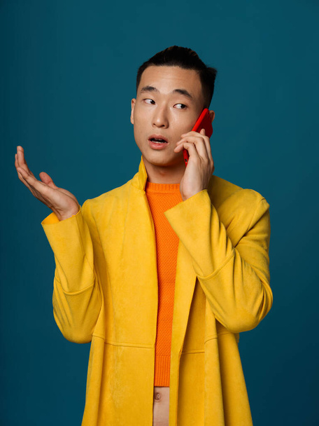 A fashionable man in a yellow jacket and an orange T-shirt is talking on the phone on a blue background - Fotoğraf, Görsel