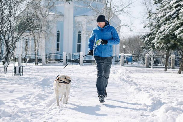 Dueño del perro con su mascota divirtiéndose en el parque en un día de invierno nevado. El hombre y un labrador recuperador corren y están activos al aire libre entre montones de nieve. Concepto de amistad y unión - Foto, Imagen