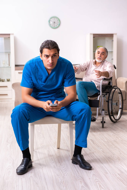 Oude man in rolstoel en jonge slechte verzorger binnen - Foto, afbeelding