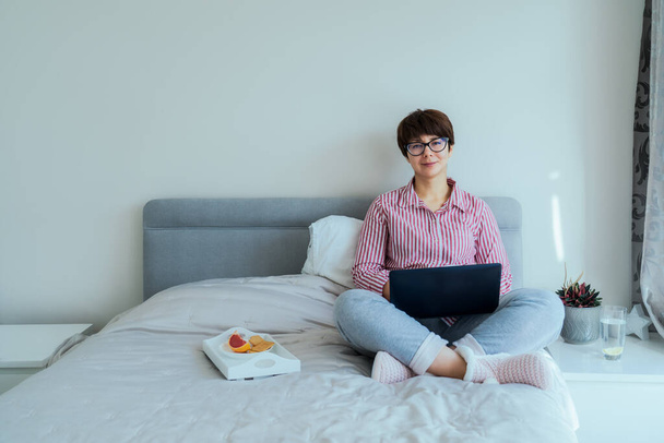 Mujer caucásica en gafas, camisa y pantalones de pijama usando portátil sentado en su cama y desayunando. Aspecto apropiado para el trabajo remoto desde el hogar durante el bloqueo. Enfoque selectivo, espacio de copia. - Foto, imagen
