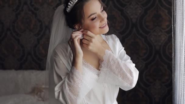 Bride in boudoir dress puts on earrings. Wedding morning preparations. Woman in night gown and veil - Footage, Video
