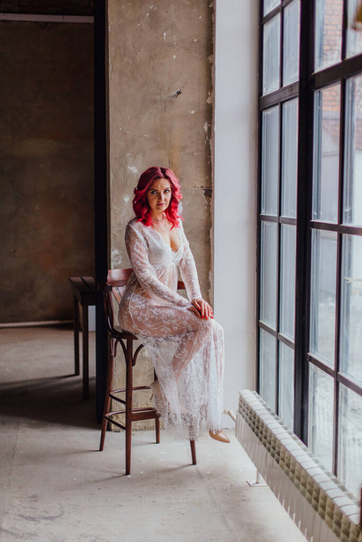 Attractive woman with pink hair in white light dress posing on the bar chair near large window. Lady in gentle lace peignoir sitting in a dark room with concrete floor and walls - Foto, Bild